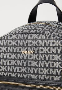 Fekete-fehér DKNY hátizsák, amely szövéssel készült anyagból készült, ismétlődő logómintával, arany zipzárral és fekete bőr részletekkel. Kerek forma.