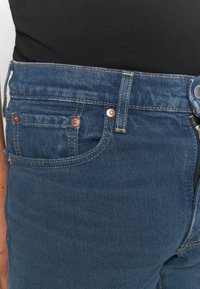 Blå denimjeans med kopparnitar, kontrastsömnad, en sidficka och dragkedja. Materialet verkar robust och texturerat.
