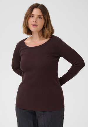 Femme aux cheveux châtains mi-longs portant un haut à manches longues marron foncé et un pantalon foncé, debout les mains derrière le dos sur un fond uni.