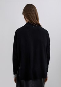 Svart, stickad tröja med hög hals, långa ärmar och lös passform. Har en liten, rund detalj nära kragen. Slät textur.