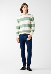 Sweter w paski w kolorze zielonym i kremowym, dzianinowa faktura, okrągły dekolt, napis "GUESS JEANS". Obcisłe niebieskie jeansy w ciemnym odcieniu, zestawione z czarnymi butami.
