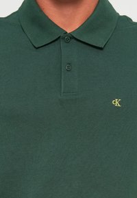 Donkergroen polo met twee losgeknoopte knopen en het gele "cK" logo geborduurd op de linkerborst.