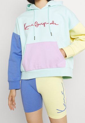 Person iført en pastel farveblok-hættetrøje med rød scripttekst og matchende blå og gule cykelshorts med en ansigtslinjetegning på det ene ben.