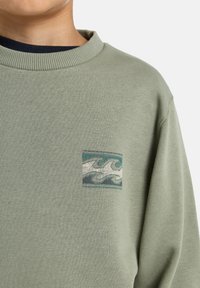 Felpa verde con collo a girocollo a coste, caratterizzata da un piccolo design di onda ricamato in blu, bianco e verde sul petto.