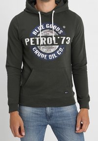 Sudadera con capucha de color verde oscuro hecha de mezcla de algodón, con bolsillo frontal, cordones blancos y un diseño gráfico redondo con texto en azul y blanco.