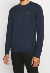 Marineblauer, gestrickter Pullover mit Rundhalsausschnitt, gerippten Bündchen und Saum. Verfügt über ein kleines, mehrfarbig gesticktes Logo auf der Brust.