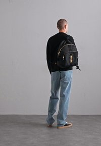Bărbat îmbrăcat cu pulover negru, blugi deschiși la culoare, și adidași negri, cu un rucsac negru, stând în fața unui perete simplu de culoare gri deschis.