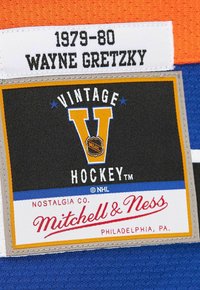 1979-80 Wayne Gretzky Namensschild über dem Mitchell & Ness Vintage Hockey-Logo auf einem orange-blauen Stoffhintergrund.