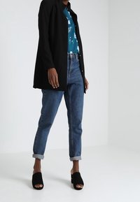 Chaqueta larga negra sobre una camisa floral verde azulado, combinada con jeans oscuros remangados y mule negro de punta abierta. Un atuendo sencillo y moderno.