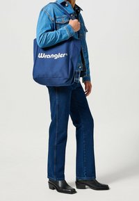 Borsa a spalla in tela blu navy con logo bianco "Wrangler", caratterizzata da manici robusti e una semplice forma rettangolare, esposta con un outfit di jeans.