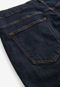 Tmavomodrá rifľovina s zadným vreckom a kontrastným oranžovým prešitím, ukazujúc textúru a detaily švov.