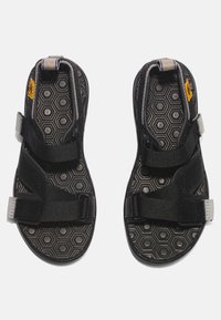 Svart sandal med justerbara remmar, texturerad hexagonal innersula och gummisula. Har grå detaljer och slitstarka nylontyg.