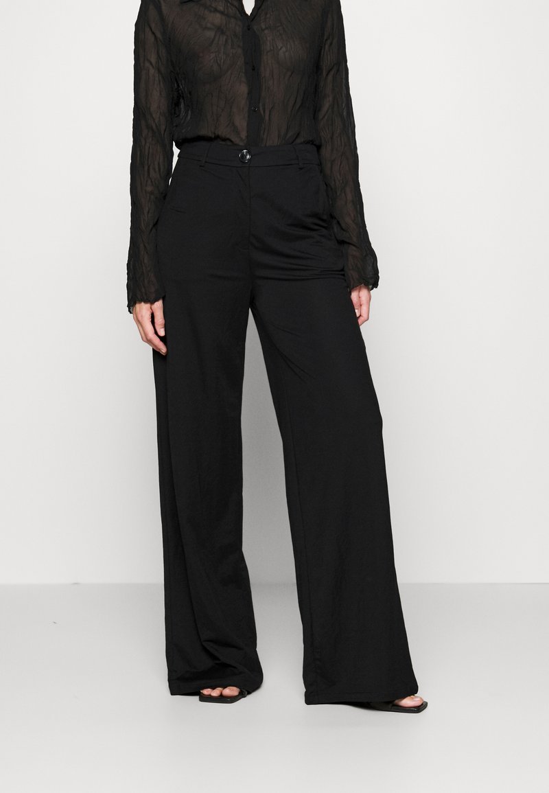 Zwarte wijde broek van soepel stof, met een hoge taille en een sluiting met één knop, gecombineerd met een transparante zwarte blouse met lange mouwen.