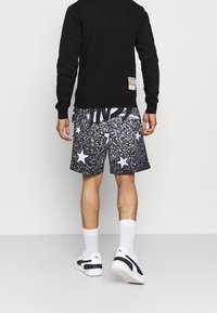 Sweat-shirt noir à col rond, associé à un short noir et blanc avec un motif moucheté et des étoiles. Chaussettes blanches et baskets noires.