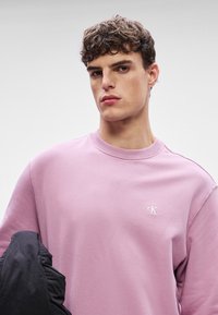 Sudadera rosa claro con cuello redondo y puños acanalados, con un pequeño logo blanco "CK" en el pecho. Chaqueta negra colgada del brazo.