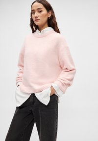 Ανοιχτό ροζ πλεκτό πουλόβερ με στρογγυλή λαιμόκοψη, χαμηλωμένους ώμους και ριμπ μανσέτες, φορεμένο πάνω από ένα λευκό πουκάμισο με γιακά και συνδυασμένο με μαύρο τζιν.