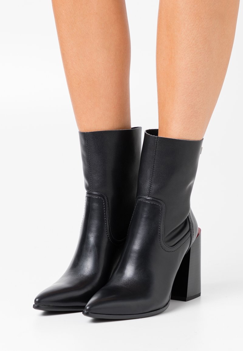 Bottes mi-mollet en cuir noir avec bouts pointus et talons hauts épais portées sur des jambes nues sur fond blanc.