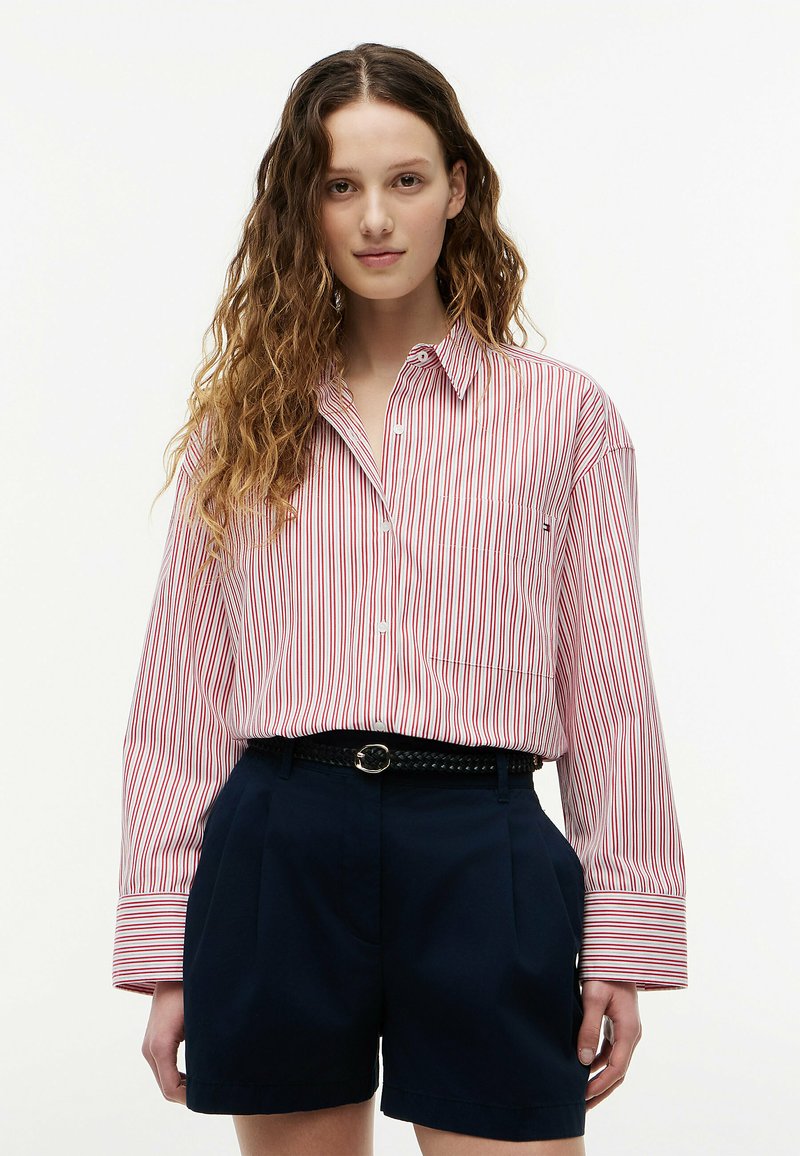 Jeune femme aux longs cheveux bouclés portant une chemise à rayures rouges et blanches à boutons, rentrée dans un short plissé bleu marine avec une ceinture noire.