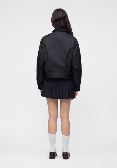 Personne debout de dos, portant une veste noire, une jupe plissée noire, des chaussettes blanches et des chaussures marron foncé sur un fond clair uni.