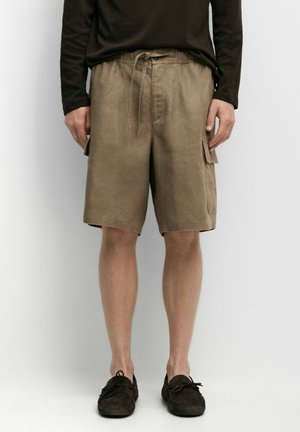 Mann trägt braunes Langarmshirt, beige kniehohe Cargoshorts mit Seitentaschen und dunkelbraune lässige Schnürschuhe, steht an einer weißen Wand.