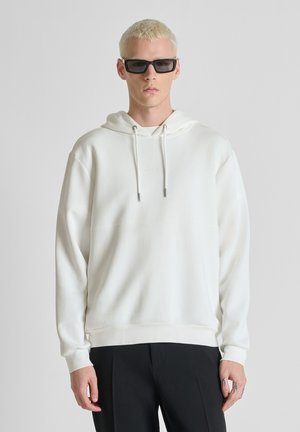 Homme blond portant des lunettes de soleil rectangulaires noires, un sweat à capuche blanc et un pantalon noir, debout devant un fond gris clair uni.