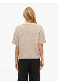 Beeži lühikeste varrukatega kampsun, mis on valmistatud pehmest, tekstureeritud kudumismaterjalist, koos lahtise lõike ja ümara kaelusega, mida kantakse tumedate pükstega.