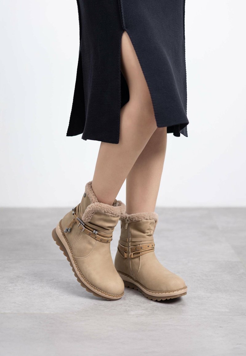 Botas de invierno marrones con forro de piel sintética, altura hasta el tobillo, correas decorativas y hebillas, y suela de goma texturizada, combinadas con un vestido negro de punto.