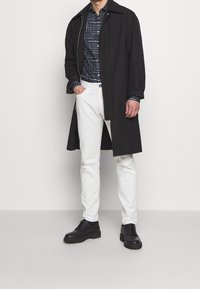 Abrigo largo negro sobre una camisa azul marino con estampado, combinado con pantalones blancos y botas negras. Cuenta con detalles de cremallera y botones.