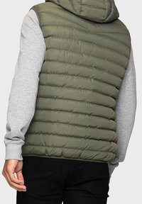 Veste matelassée sans manches vert olive avec capuche, texture matelassée, et un t-shirt à manches longues gris en dessous. Pantalon noir partiellement visible.