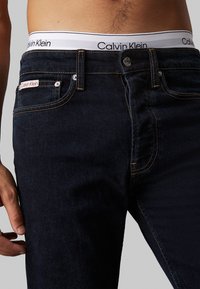 Jeans en denim bleu foncé avec une coupe slim, des coutures orange contrastées et une taille blanche Calvin Klein avec un détail de logo.