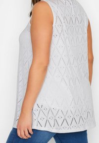 Ärmlös vit topp med diamantformad hålmönster, bärs med blå jeans, visas från sidan och bakifrån på en person som står.