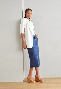 Weißer Blazer über einem weißen Top, kombiniert mit einem Jeans-Bleistiftrock und orangefarbenen Keilsandalen mit Korkplattform, stehend gegen eine Wand.