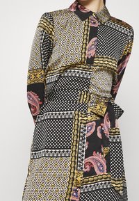 Sukienka z długim rękawem i kołnierzykiem, która łączy wzory paisley, kropki oraz geometryczne w kolorach czarnym, białym, złotym i czerwonym. W talii ozdobny detal w postaci związania.