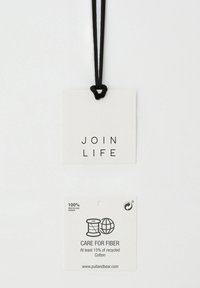 Vitt pappers hängtag med svart text "JOIN LIFE" och information om material, återvunnen innehåll och skötselråd längst ner.