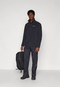 Črni pulover iz flisa s četrtinskim zadrgo in izvezenim logotipom, v kombinaciji s črnimi hlačami in črnim nahrbtnikom. Tkanina z teksturo.