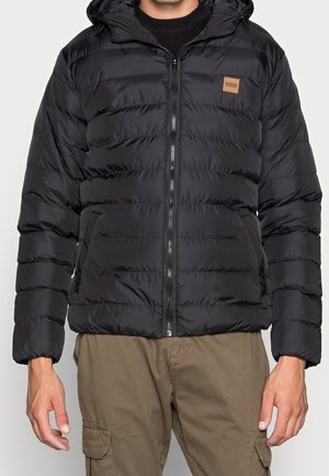 Homme portant une veste matelassée noire à capuche avec fermeture éclair à l'avant et un pantalon cargo marron, debout devant un fond clair uni.
