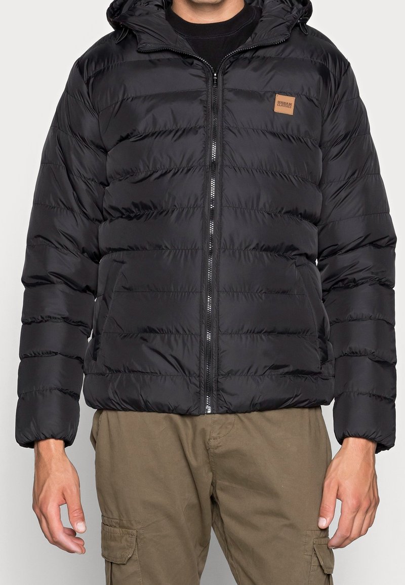 Homme portant une veste matelassée noire à capuche avec fermeture éclair à l'avant et un pantalon cargo marron, debout devant un fond clair uni.