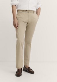 Beige Baumwoll-Chinos mit geradem Bein, ausgestattet mit Seitentaschen und einem Knopfverschluss. Getragen mit braunen Mokassins.