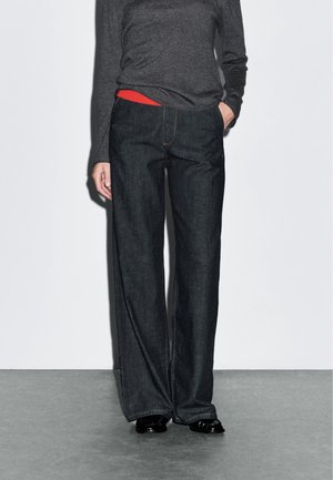 Personne portant un jean large foncé, un haut à manches longues gris foncé, une chemise rouge qui dépasse et des chaussures noires, debout sur un sol gris.