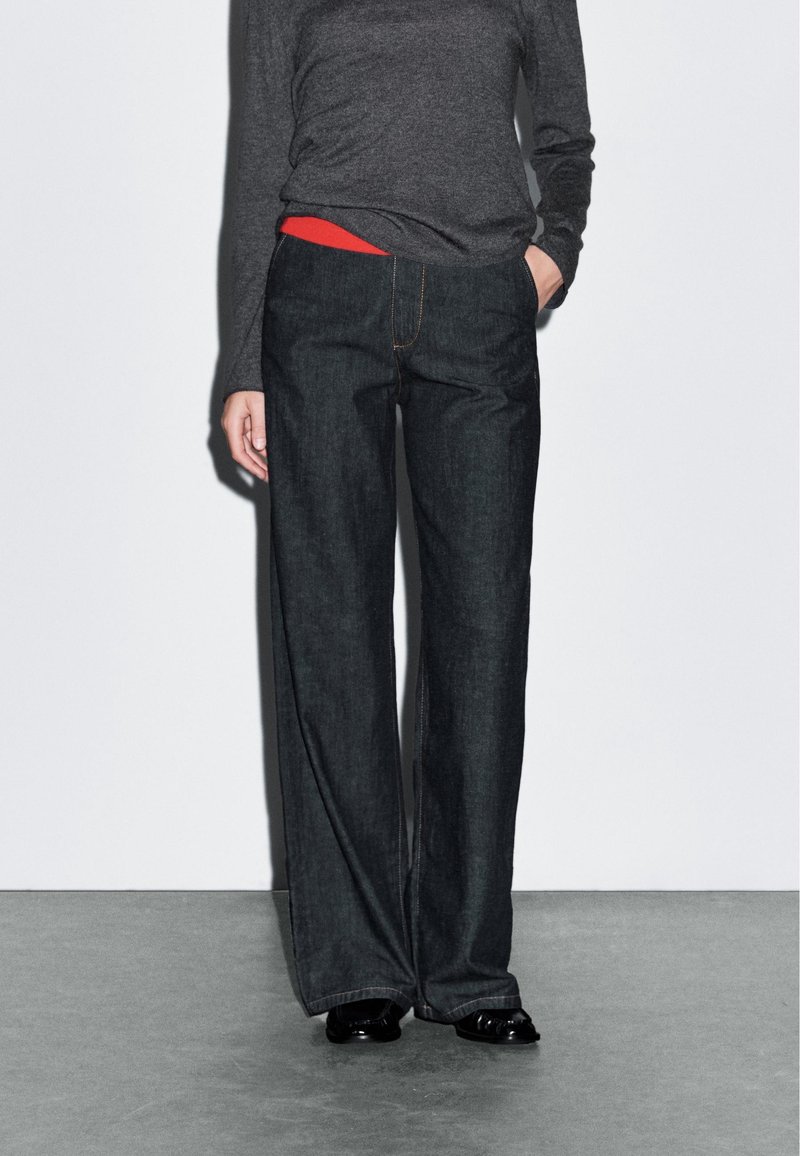 Personne portant un jean large foncé, un haut à manches longues gris foncé, une chemise rouge qui dépasse et des chaussures noires, debout sur un sol gris.