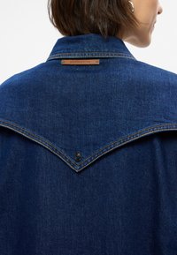 Τζάκετ τζιν σε σκούρο μπλε χρώμα, με σχέδιο μυτερού yoke, αντίθετη πορτοκαλί ραφή και δερμάτινη ετικέτα μάρκας στον γιακά.