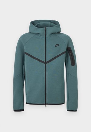 Φούτερ Nike με πλήρη φερμουάρ σε τιρκουάζ χρώμα, με μαύρη λωρίδα σε σχήμα V, τσέπη με φερμουάρ στο αριστερό μανίκι και μαύρα τερματικά φερμουάρ, εμφανιζόμενο σε λευκό φόντο.