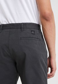 Šedé lehké kalhoty s hladkou texturou, vybavené jedním zadním kapsou a malou černou štítkem s bílým textem.