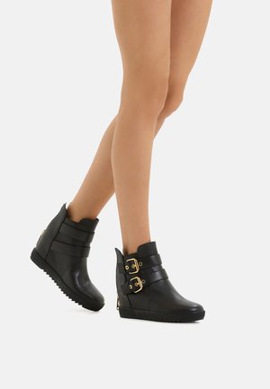 Schwarze Keil-Ankleboots aus glattem Material, mit zwei goldenen Schnallen und strukturiertem Sohlen für besseren Halt. Verstellbares Riemendesign.