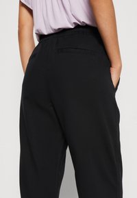 Pantalons noir à taille élastique avec poches latérales, texture lisse, et une zone des hanches ajustée, se rétrécissant vers les jambes.