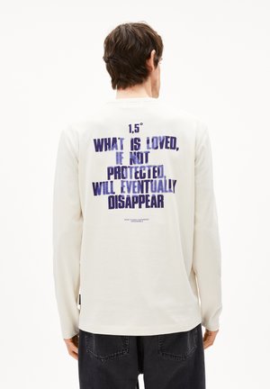 Langärmliges cremefarbenes Shirt mit lockerem Schnitt, das auf der Rückseite blauen Text zeigt: "1,5° WAS GELIEBT WIRD, WENN NICHT GESCHÜTZT, WIRD SCHLIEßLICH VERSCHWINDEN."