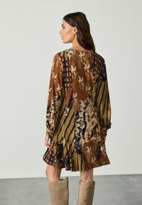 Abito a maniche lunghe in toni terrosi con motivi astratti, caratterizzato da una vestibilità comoda, gonna plissettata e scollo rotondo, abbinato a stivali chiari.