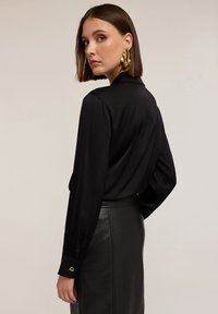 Zwarte satijnen blouse met lange mouwen en een kraag, gecombineerd met een zwarte leren rok. Accentdetail op de mouwmanchet. Model van achteren weergegeven.