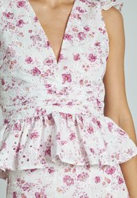 Top peplum floreale bianco con fiori rosa, dotato di scollatura a V profonda e dettagli in occhialini. Tessuto leggero e texturizzato con rouches.