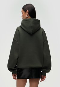 Felpa oversize verde oliva con un ampio cappuccio e spalle scese, abbinata a una mini gonna nera lucida. Tessuto morbido, vestibilità comoda.